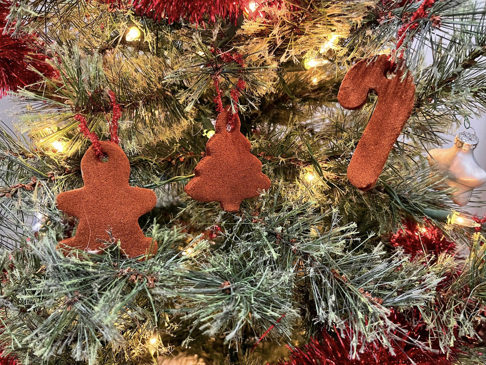 Cinnamon dough ornaments in the shape of a gingerbread man, a Christmas tree, and a candy cane hanging on a Christmas tree with red ribbon and white lights in the background.